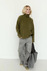 A woman stands against a plain white wall wearing an oversized olive green turtleneck sweater, wide-leg gray jeans, and tan shoes, while holding a large dark gray tote bag.