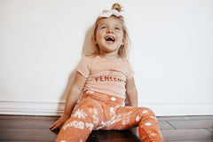 Child wearing a pink shirt with 'Penthouse' text and orange pants with white patterns, sitting on a wooden floor.