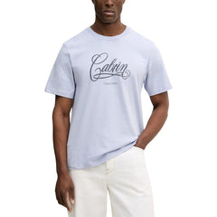 Image 1 : T-shirt homme Calvin Klein Jeans bleu lavande avec logo script 'Calvin' imprimé sur la poitrine, porté avec un jean blanc — Delvale