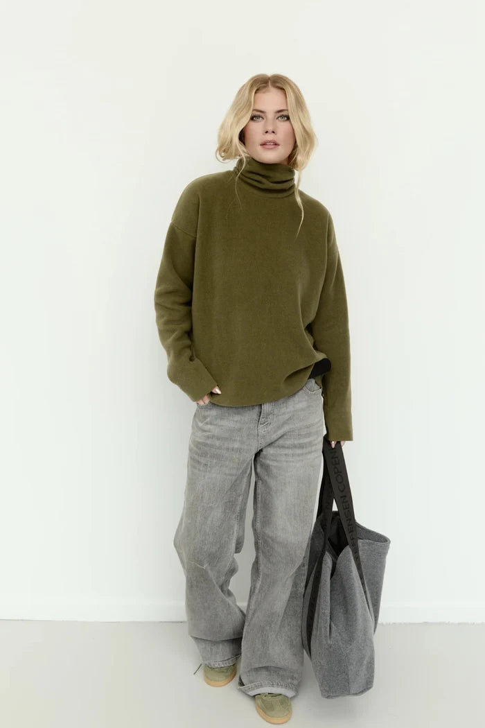 A blonde model stands against a plain white studio wall wearing an oversized olive green turtleneck sweater, wide-leg gray jeans, and beige shoes, while holding a large black tote bag.
