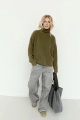 A woman stands against a plain white wall wearing an olive green turtleneck sweater, wide-leg light gray pants, and olive shoes, while holding a large gray tote bag.